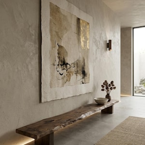 May include: A large rectangular wall art piece featuring abstract beige, brown, and gold leaf accents on textured paper, mounted above a long, dark brown live-edge wooden bench with block legs, positioned in a room with light-colored walls.