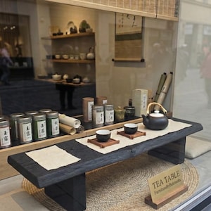 Op de afbeelding: Een theetafeldisplay in een etalage. De tafel is van donker hout met een jute vloerkleed eronder. Theekopjes, een theepot en theebussen zijn op de tafel geplaatst. Het bordje luidt "TEA TABLE FurnitureByflukmedis."