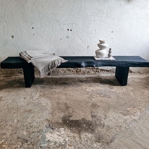 May include: A dark stained wooden bench with a natural edge, with a grey fringed blanket and a white ceramic sculpture. The bench is supported by two black rectangular legs. A small decorative item sits on a square tile.
