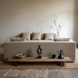 Könnte beinhalten: Ein beiges Polstersofa mit fünf passenden quadratischen Kissen, das hinter einer niedrigen, rechteckigen Holzbank steht. Auf der Bank befinden sich eine flache Schale mit grauen Steinen, eine dunkle abstrakte Skulptur, eine kleine schwarze Schale und eine schwarze Teekanne.