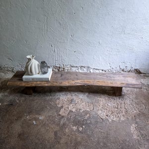 Könnte beinhalten: Rustikale Holzbank mit natürlicher Kante, auf der eine weiße Vase, ein kleines Glasgefäß und ein Buch stehen. Die Bank wird von zwei kurzen Holzbeinen getragen. Die Gegenstände sind vor einer strukturierten, hellgrauen Wand angeordnet.