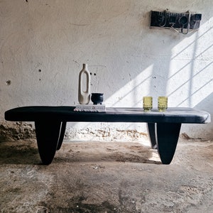 Könnte beinhalten: Ein schwarzer Holz-Couchtisch mit einem einzigartigen, skulpturalen Design. Der Tisch hat eine rechteckige Platte und zwei geschwungene Beine auf jeder Seite. Auf dem Tisch stehen zwei grüne Gläser, eine weiße Keramikvase und ein kleiner schwarzer Keramiktopf.