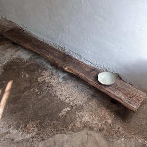 May include: A long, rectangular wooden bench with a dark brown, weathered grain and visible knots. The bench features a low-profile design supported by short legs. A single, small, light-colored ceramic bowl sits on the right side of the surface.