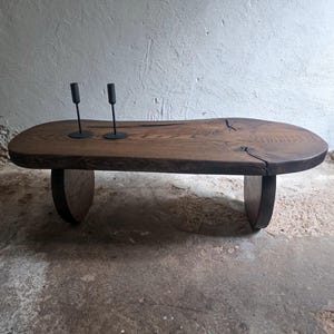 Könnte beinhalten: Ein niedriger Couchtisch aus einer massiven, dunkelbraunen Holzplatte mit natürlicher Baumkante und sichtbarer Maserung. Der Tisch steht auf zwei geschwungenen Holzfüßen und trägt zwei schlanke, schwarze Kerzenhalter aus Metall.