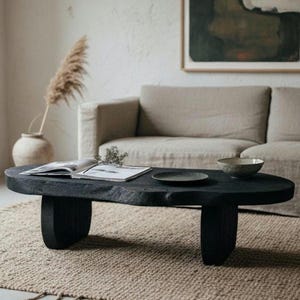 May include: A low-profile coffee table crafted from a single piece of dark, charred wood with a natural, irregular live edge. The table features two thick, sturdy slab legs and a smooth, matte black finish, providing a solid surface for decor.