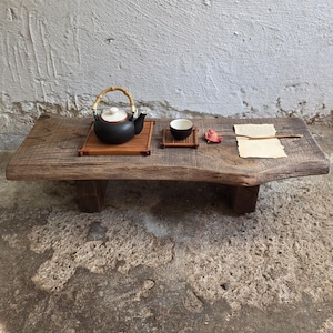 Puede incluir: Una mesa baja rectangular de madera con borde natural, que presenta una tetera de cerámica negra con asa de bambú sobre una esterilla de bambú, una pequeña taza de cerámica negra sobre un posavasos de madera, una flor seca y un papel pergamino con texto.