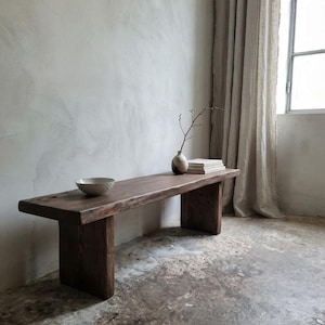 Pode incluir: Um banco de madeira castanho-escuro com um assento retangular espesso e duas pernas de placa resistentes. No banco, encontram-se uma pequena taça texturizada de cor clara, um vaso de cerâmica bicolor com um ramo fino e livros empilhados.