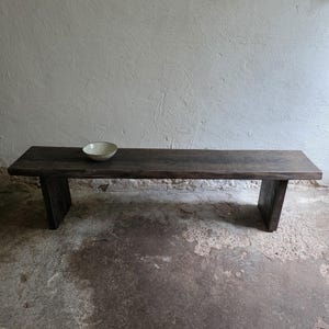 May include: A long, rectangular dark brown wooden bench with a flat top and two solid vertical slab legs. A small, shallow, light-coloured speckled bowl sits on the left side of the bench surface. The wood grain is visible across the entire bench.