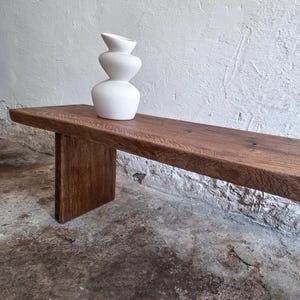 May include: A white ceramic vase with a stacked, organic, three-tiered shape sits on a long, rectangular wooden bench. The bench features a dark brown wood grain finish and is supported by a thick, flat vertical leg on the left side.