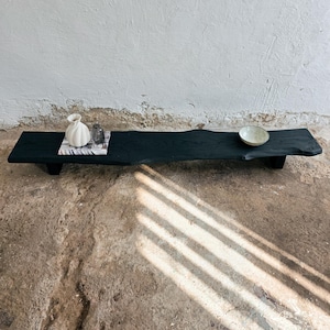 Könnte beinhalten: Ein langes, dunkel gebeiztes Holzserviertablett mit natürlicher Kante. Das Tablett enthält eine kleine weiße Vase, eine klare Glasflasche und eine kleine, hellfarbige Schale. Das Tablett steht auf kurzen Beinen.