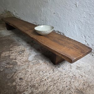 Könnte beinhalten: Eine lange, niedrige Holzbank mit natürlicher Baumkante und dunkelbraunem Finish, die auf zwei Blockbeinen ruht. Eine einzelne, runde, cremeweiße Keramikschale mit einer texturierten, genoppten Oberfläche steht in der Mitte der Bank.