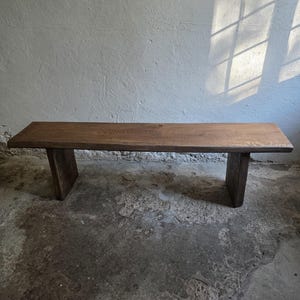 Op de afbeelding: Een rechthoekige houten bank met een platte, horizontale zitting die wordt ondersteund door twee verticale paneelpoten. De bank is gemaakt van donkerbruin hout met een zichtbare houtnerf en een glad afgewerkt oppervlak.