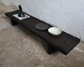 May include: A dark stained wooden bench with a rectangular top and two supporting legs. A black vase, a black plate, and a white bowl are arranged on the bench. The bench is set against a white wall and a concrete floor.