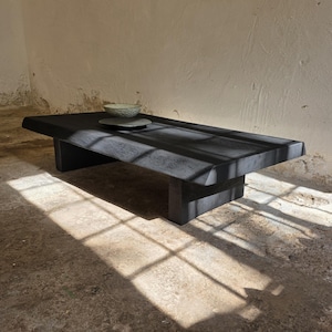 Op de afbeelding: Een lage, rechthoekige salontafel met een matzwarte afwerking en een dik, massief houten blad. De tafel heeft twee stevige blokpoten en een zichtbare houtnerfstructuur. Twee kleine, neutraal gekleurde keramische kommen staan op het oppervlak.