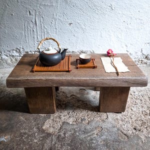 Op de afbeelding: Een lage rechthoekige houten tafel met een donkere matte afwerking. Op de tafel staat een zwarte theepot met een wit deksel en bamboe handvat op een bamboemat, een klein bijpassend theekopje op een onderzetter en een gedroogde roze roos op papier.