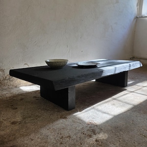 May include: A low-profile, rectangular coffee table crafted from solid, dark-stained wood with a thick, natural-edge slab top and two sturdy block legs. A light-colored, textured ceramic bowl and a small, flat metallic plate sit on the surface.
