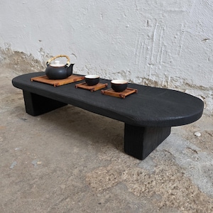 Puede incluir: Una mesa de té baja de madera negra con una parte superior redondeada y dos patas cortas. Una tetera negra con asa de bambú se encuentra sobre una esterilla de bambú, junto con dos tazas de té pequeñas sobre esterillas individuales. La mesa mide unos 100 cm de largo.