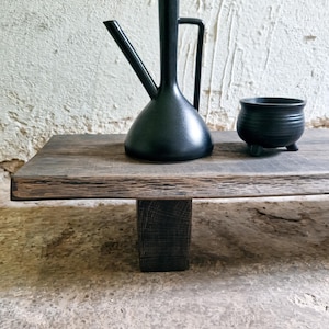 Op de afbeelding: Een matzwarte keramische oliekruik met een lange tuit en handvat, geplaatst naast een kleine, ronde, driepotige zwarte keramische kom. Beide items staan op een rechthoekige houten plank met een donkere, getextureerde afwerking en één steun.