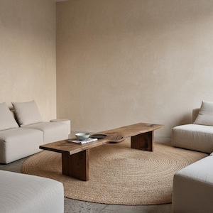 Op de afbeelding: Een moderne woonkamer met neutrale tinten. Een grote, donkere houten salontafel staat op een rond jute vloerkleed. Beige banken en stoelen omringen de tafel. Een kom en een bord staan op de tafel. De muren zijn lichtbeige.