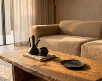 May include: A rectangular wooden coffee table with a natural live edge, featuring a dark brown finish and sturdy block legs. On the surface sits a black ceramic pitcher, a small black bowl, a book, and a round black textured plate.
