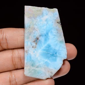 May include: A polished, irregular-shaped piece of larimar gemstone. The stone displays a mix of light blue and white hues with darker blue veining and some brown inclusions. The stone is held in a hand against a black background.