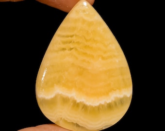 Puede incluir: Una gema pulida en forma de lágrima, de color amarillo vibrante y con un patrón en capas y bandas. La piedra tiene una superficie brillante y se sostiene sobre un fondo negro. El color de la gema varía de amarillo claro a un tono dorado más intenso.