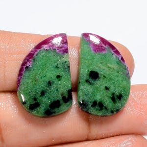 Puede incluir: Un par de cabujones de piedra preciosa verde y rosa con motas negras. Los cabujones tienen forma de lágrima y una superficie lisa y pulida.