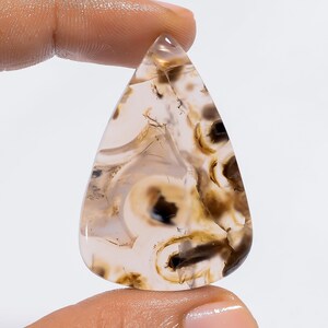 Puede incluir: Un cabuj&oacute;n de &aacute;gata en forma de l&aacute;grima con un patr&oacute;n de remolino de marr&oacute;n, blanco y negro. La piedra es transl&uacute;cida y tiene una superficie lisa y pulida.