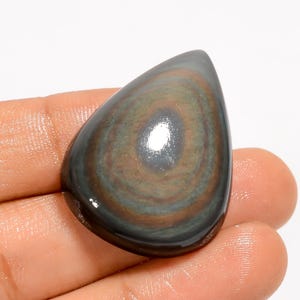 Puede incluir: Una gema pulida en forma de l&aacute;grima con un patr&oacute;n arremolinado de tonos marrones, grises y verdes. La piedra tiene una superficie brillante y se sostiene en una mano, mostrando su belleza natural y sus variaciones de color &uacute;nicas.