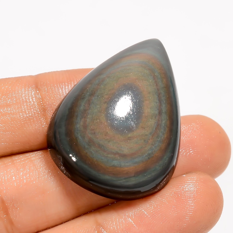 Puede incluir: Una gema pulida en forma de l&aacute;grima con un patr&oacute;n arremolinado de tonos marrones, grises y verdes. La piedra tiene una superficie brillante y se sostiene en una mano, mostrando su belleza natural y sus variaciones de color &uacute;nicas.