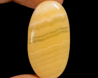 May include: Oval-shaped gemstone with a polished surface. The stone features a blend of yellow and white hues, with subtle darker bands running across its length. The gemstone is held between two fingers against a black background.