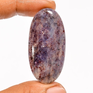 Puede incluir: Una piedra preciosa ovalada con superficie pulida. La piedra presenta una mezcla de tonos morados, rojos y blancos, con un patrón moteado. La piedra preciosa se sostiene entre dos dedos.