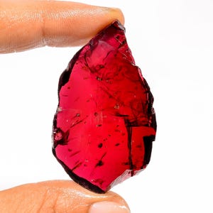 May include: A raw, uncut, deep red gemstone with a translucent quality. The irregularly shaped stone is held between two fingers against a white background, showcasing its natural, rough texture and rich color.