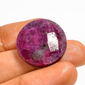 Peut inclure: Une pierre précieuse ronde et polie, d'un magenta profond, parsemée d'inclusions roses et grises plus claires. La gemme est tenue dans une main, mettant en valeur sa surface lisse et réfléchissante. La forme de la pierre est un cercle parfait.