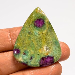 Puede incluir: Una gema pulida con forma de lágrima. La piedra presenta una base verde con inclusiones de color magenta vibrante. La gema se sostiene en una mano, mostrando sus patrones naturales y variaciones de color únicas.