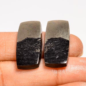 May include: Two rectangular pyrite stones with a polished, metallic appearance. The top portion of each stone is a muted silver color, transitioning to a textured, dark charcoal base. The stones are held in a hand against a white background.