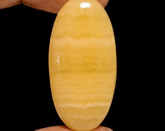 Puede incluir: Una gema pulida de forma ovalada con un tono amarillo cálido y sutiles bandas horizontales. La piedra se sostiene entre dos dedos sobre un fondo negro, resaltando su superficie lisa y brillante. El color de la gema varía de amarillo claro a medio.
