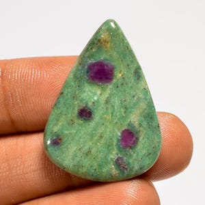 May include: A teardrop-shaped gemstone with a green base color and several deep red inclusions. The stone has a polished surface and is held by a hand. The gemstone is likely a type of ruby in zoisite.