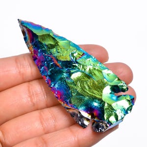 Puede incluir: Una punta de flecha iridiscente con un esquema de colores verde, azul y morado. La punta de flecha está hecha de un material liso y pulido.
