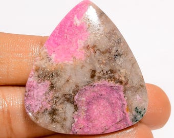 May include: A polished gemstone with a triangular shape. The stone features a blend of pink and gray hues, with areas of vibrant pink and speckled gray. The gemstone is held in a hand, showcasing its smooth, reflective surface.