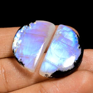 May include: Two polished moonstone cabochons, each a semi-circle shape. The stones display a play of color with blue, white, and lavender hues. The edges are outlined with a black border. The gemstones are displayed against a dark background.