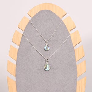 May include: Two silver necklaces with iridescent abalone shell pendants. The top necklace has a round pendant and the bottom necklace has a teardrop pendant. The necklaces are displayed on a wooden stand.
