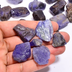 May include: A collection of raw, rough-cut, purple-blue gemstones. The stones are various sizes and shapes, with some having a more polished look.