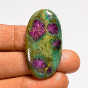 May include: Oval-shaped gemstone with a vibrant mix of colors. The stone features a green base with patches of yellow and deep magenta-pink inclusions. The polished surface reflects light, highlighting the natural patterns and textures of the stone.