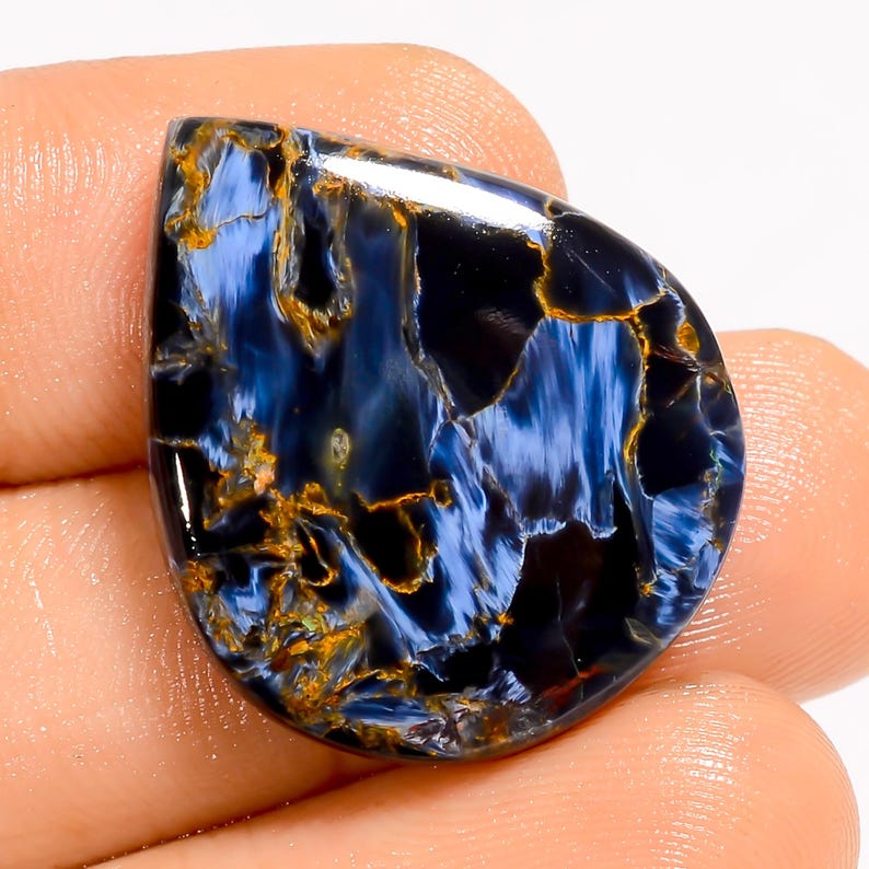 May include: A polished, teardrop-shaped gemstone with a striking pattern of deep blues, blacks, and golden-brown veins. The stone's surface is smooth and reflective, showcasing a natural, swirling design. The gemstone is held in a hand.