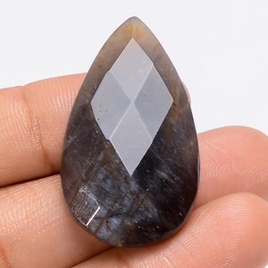 May include: A pear-shaped cabochon gemstone with a diamond-shaped pattern in the center. The gemstone is a dark gray color with a hint of blue.