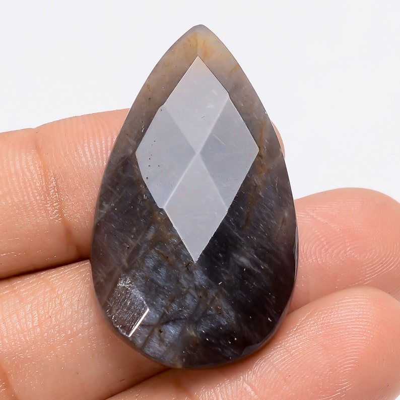 May include: A pear-shaped cabochon gemstone with a diamond-shaped pattern in the center. The gemstone is a dark gray color with a hint of blue.