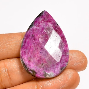 May include: A faceted, teardrop-shaped gemstone with a vibrant pink hue, accented by darker gray and black inclusions. The polished surface reflects light, showcasing the stone's internal structure. The gem is held in a hand, highlighting its size and shape.