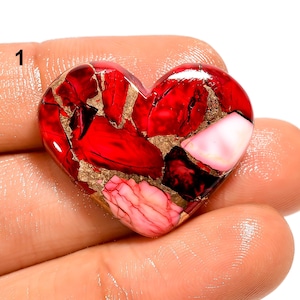 Peut inclure: Une pierre précieuse en forme de cœur, principalement rouge avec des inclusions dorées et roses. La surface polie reflète la lumière, soulignant les motifs uniques de la pierre. Le chiffre "1" est visible dans le coin supérieur gauche.