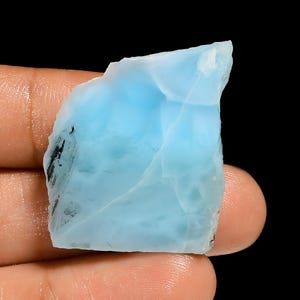 May include: A rough-cut, light blue gemstone with a translucent appearance. The stone has a natural, irregular shape and a smooth, polished surface. The gemstone is held against a black background.